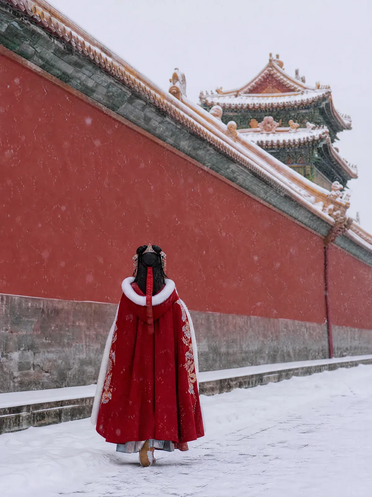 A woman in a red hanfu cape walking past the snowy red walls of the Forbidden City